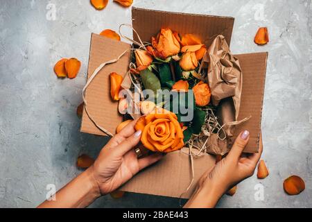 Die Hände der Frau halten offene Kiste mit orangefarbenen Rosen darin, Nahaufnahme. Geschenkkonzept. Weißer Zementhintergrund mit Rosenblättern, Draufsicht. Stockfoto