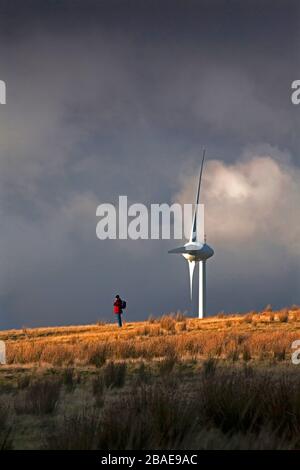 Einsamer Bergwanderer auf dem Hügelkamm mit Windturbine und stürmischem Himmel mit dramatischem Licht, Wales, Großbritannien Stockfoto