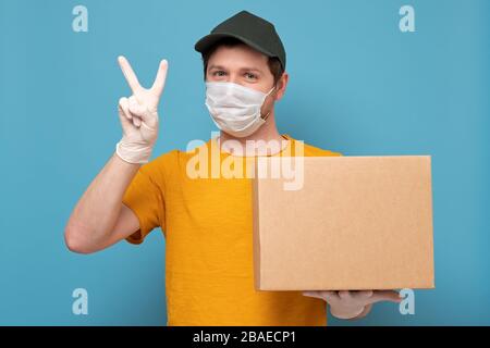 Junger Auslieferer in medizinischer Maske, der zwei Finger zeigt, die eine auf weißem Hintergrund isolierte cardbox halten und tragen Stockfoto