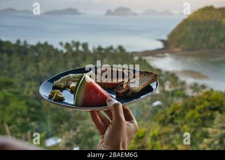 Mann mit gesundem kontinentalen Frühstück vor einem paradiesischen Strand mit Blick auf El Nido, Philippinen. Wassermelone, Rührei, Toast und Butter Stockfoto