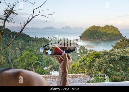 Mann mit gesundem kontinentalen Frühstück vor einem paradiesischen Strand mit Blick auf El Nido, Philippinen. Wassermelone, Rührei, Toast und Butter Stockfoto