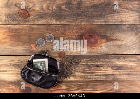 Offene Tasche aus schwarzem Leder mit Münzen, einem Cent, einem Viertel-Dollar und einer Banknote von 2 Dollar in der Nähe. Finanzkrise, Armut, Geldmangel. Auf Holz B. Stockfoto