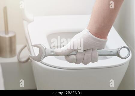 Klempnerhand mit Schraubenschlüssel auf dem hintergrund des toilettensitzes Nahansicht Stockfoto