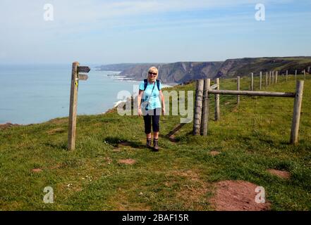 Lone Woman Hiker Walking on Clifftop by Wooden Signpost zwischen Welcome Mouth & Spekes Mill auf dem South West Coast Path, North Devon. England, Großbritannien. Stockfoto