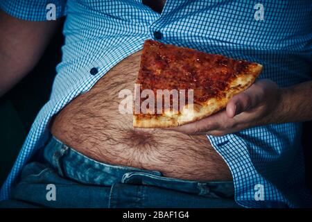 Nahaufnahme eines kaukasischen Mannes, mit einem haarigen Bierbauch, auf dem Sofa sitzend, Pizza essen Stockfoto