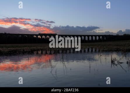 Der Zug der nördlichen Züge der Klasse 144 mit Schrittmacherzug, der den bogenförmigen Penistone-Viadukt überquert und eine SonnenuntergangsSilhouette in einem überfluteten Feld reflektiert Stockfoto