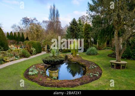 Landhausgarten (Teichwasserspiegelungen, Rundsitz, dekorative Urne, Schotterweg, Topiary, Rasen, reife Bäume) - York Gate Garden, Leeds UK. Stockfoto
