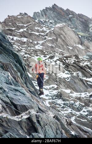 Volle Länge des männlichen Alpinisten in Sonnenbrille und Sicherheitshelm aufsteigenden hohen Berg. Mann Kletterer hält fest Seil und schaut nach unten. Konzept des Bergsteigens, Alpinismus und Klettern. Stockfoto