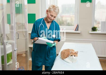 Reife Frau in Uniform macht Notizen in der Krankenkarte verschreibt sie ein Medikament für Hauskatze Stockfoto
