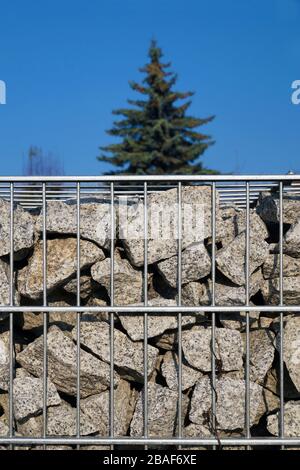 Nahaufnahme auf einem mit Granit-Splittern gefüllten Gabion. Himmel und Kiefer im Hintergrund. Stockfoto