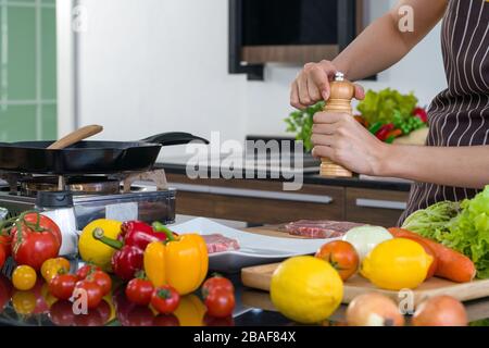 Junge Hausfrau legt Pfeffer auf das Schweinefleisch, bevor sie es zu einem Steak einkocht. Morgendliche Atmosphäre in einer modernen Küche. Die Küchenecke voll Variou Stockfoto