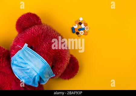 Einsamer Roter Teddybär in einer schützenden medizinischen Maske auf gelbem Hintergrund mit Atemmasken und Pillen. Coronavirus Kovid-19-Präventionskonzept. Stockfoto