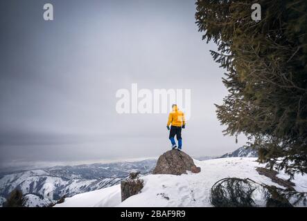 Tourist in gelben Jacke steht an der Spitze des Felsens in den verschneiten Bergen Stockfoto