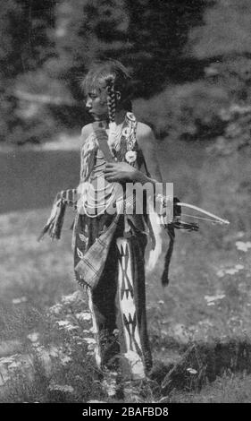 American Indian / Indianer Junge posiert mit Bogen & Pfeile in einer Scheide oder Köcher auf dem Rücken, um 1907. Er trägt eine feine Sammlung von wunderschön gestalteten und Perlen Kleidung einschließlich Leggings, ein Breechcloth, Mokassins (nicht sichtbar) & Strings von Perlen um seinen Hals. Er sieht nicht glücklich aus. Tatsächlich ließ der weiße Fotograf, wie es damals üblich war, den Jungen all die authentische, aber nicht alltägliche Kleidung anziehen, um nur zu posieren, damit der Fotograf ein romantisiertes indisches Bild an Touristen verkaufen konnte. Um meine anderen Vintage-Bilder zu sehen, Suche: Prestor Vintage West Stockfoto