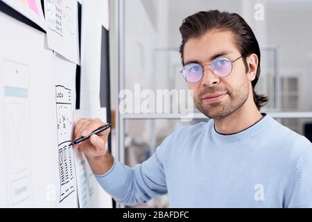 Portrait des gutaussehenden UI-Designers in runder Brille mit App-Schema auf dem Planungsbrett im Büro Stockfoto