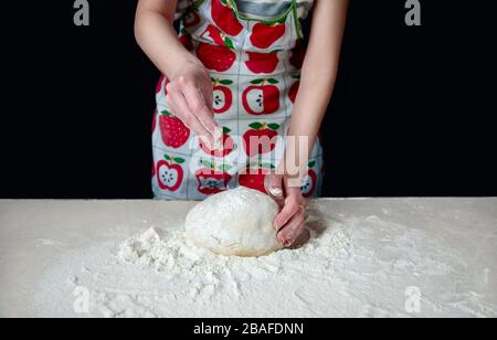 Frau übergibt Knäuel den Teig und streut ihn mit weißem Mehl auf den Küchentisch auf dunklem Hintergrund. Stockfoto