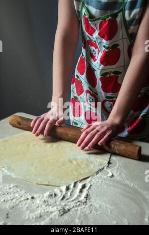 Frau rollt Teig Rollstift auf dem Küchentisch auf dunklem Hintergrund. Das Strahllicht fällt im Dunkeln auf den Tisch. Stockfoto