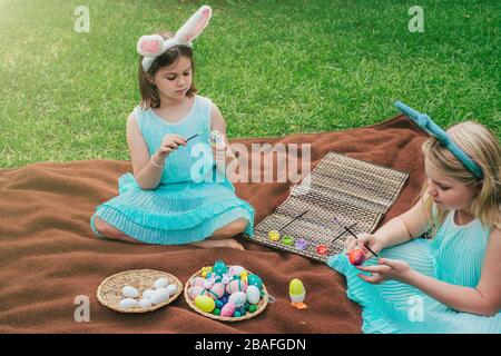 Niedliche kleine Mädchen, die am Ostertag auf der Decke auf Gras im Garten sitzen und auf den Eiern malen, tragen bunte Ohren. Stockfoto