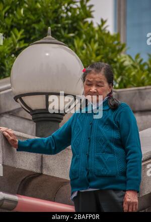 Shanghai, China - 4. Mai 2010: Ältere Frau in blauer Weste hält sich fest an der grauen Steinwand. Große Kugellaterne und grünes Laub im Rücken. Stockfoto