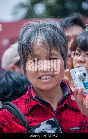 Shanghai, China - 4. Mai 2010: Frauenporträt mit kurzen, grauenden Haaren und roter Weste hält silberne Kamera, zeigt ihre Zähne und in einer trübenden Stimmung. Stockfoto