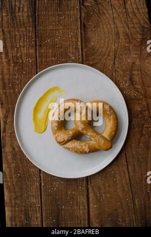 Handgemachte weiche Brezel mit Salz auf einem weißen Teller mit einem Abstrich Senf auf einem hölzernen Hintergrund. Stockfoto