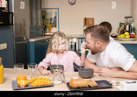 Süße kleine Tochter mit langen blonden Haaren, die ihren Vater mit Müsli füttern, während sie sowohl am Tisch sitzen als auch frühstücken Stockfoto