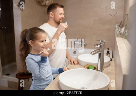 Süßes kleines Mädchen und ihr glücklicher junger Vater mit Zahnbürsten, die Zähne putzen, während sie im Badezimmer vor dem Spiegel stehen Stockfoto