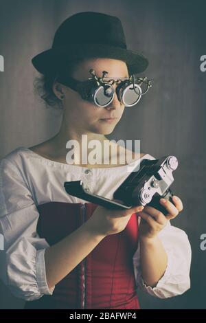 Attraktives Steampunk-Mädchen, das Brille und Hut trägt, hält die alte Kamera Stockfoto