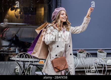 Taxi, ich bin hier! Seitenansicht der schönen blonden Frau in der Liege, die Einkaufstaschen in der Hand hält und lächelt, während sie nach dem Taxi ruft. Warten auf Kabine nach Stockfoto