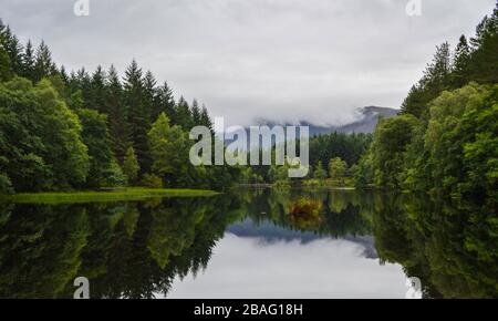 Landschaft und Seespiegelungen umgeben von einem Wald in Glencoe Lochan an einem bewölkten Tag, in den schottischen Highlands, Schottland. Stockfoto