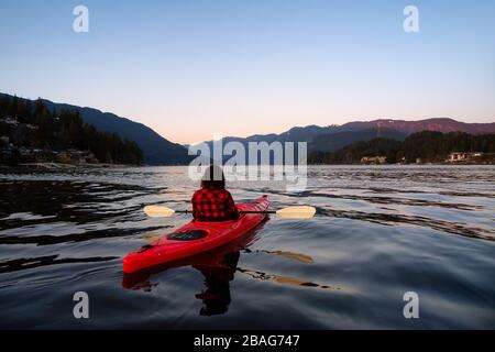 Abenteuerliches Mädchen paddeln auf einem hellen roten Kajak in ruhigem Meerwasser bei einem lebhaften und farbenfrohen Sonnenuntergang. Aufgenommen in Indian Arm, Deep Cove, North Vancouver Stockfoto