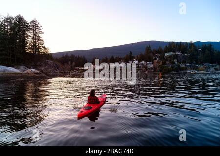 Abenteuerliches Mädchen paddeln auf einem hellen roten Kajak in ruhigem Meerwasser bei einem lebhaften und farbenfrohen Sonnenuntergang. Aufgenommen in Indian Arm, Deep Cove, North Vancouver Stockfoto