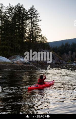 Abenteuerliches Mädchen paddeln auf einem hellen roten Kajak in ruhigem Meerwasser bei einem lebhaften und farbenfrohen Sonnenuntergang. Aufgenommen in Indian Arm, Deep Cove, North Vancouver Stockfoto