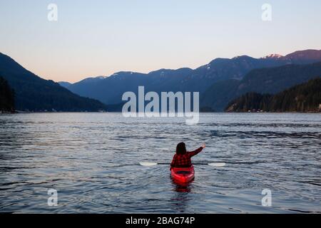 Abenteuerliches Mädchen paddeln auf einem hellen roten Kajak in ruhigem Meerwasser bei einem lebhaften und farbenfrohen Sonnenuntergang. Aufgenommen in Indian Arm, Deep Cove, North Vancouver Stockfoto