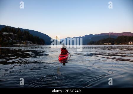 Abenteuerliches Mädchen paddeln auf einem hellen roten Kajak in ruhigem Meerwasser bei einem lebhaften und farbenfrohen Sonnenuntergang. Aufgenommen in Indian Arm, Deep Cove, North Vancouver Stockfoto