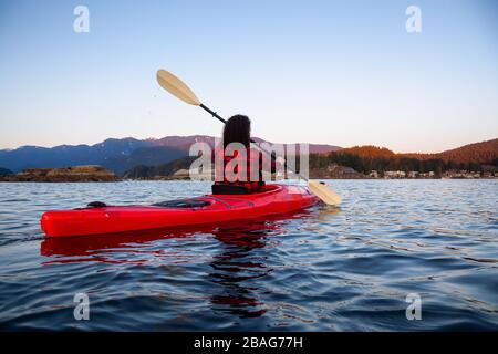 Abenteuerliches Mädchen paddeln auf einem hellen roten Kajak in ruhigem Meerwasser bei einem lebhaften und farbenfrohen Sonnenuntergang. Aufgenommen in Indian Arm, Deep Cove, North Vancouver Stockfoto