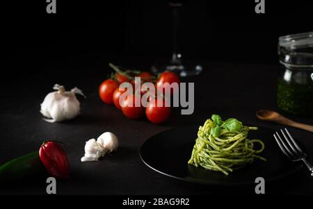 Spaghetti mit Pestosoße mit grüner Genuesauce mit frischen Tomaten auf schwarzem Teller und schwarzem Hintergrund mit Seitenlicht Stockfoto