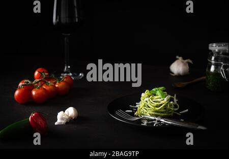 Spaghetti mit Pestosoße mit grüner Genuesauce mit frischen Tomaten auf schwarzem Teller und schwarzem Hintergrund mit Seitenlicht Stockfoto