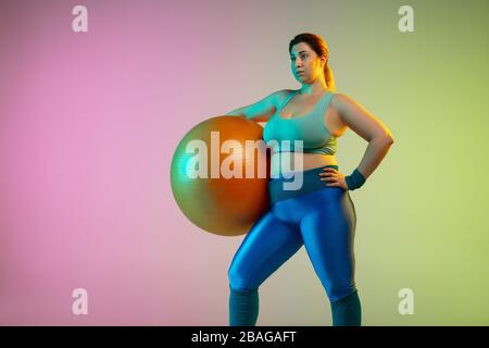 Junge kaukasische plus Größe weibliche Modell Training auf Gradient violett grünen Hintergrund in Neonlicht. Übungen mit Fitball machen. Sportkonzept, gesunder Lebensstil, körperpositiv, Gleichberechtigung. Stockfoto