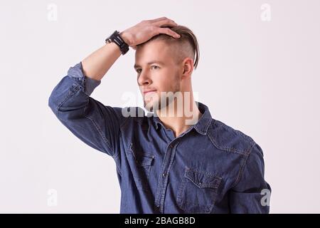 Porträt eines jungen bärtigen Hüftmännchen, der Haare berührt und wegschaut, während er vor weißem Hintergrund steht. Schöner und stilvoller Mann mit modernem Hai Stockfoto