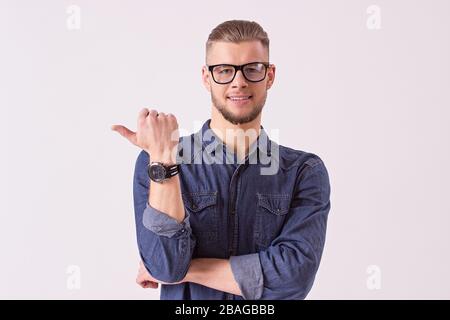 Porträt eines stylischen Mannes in Brillen, die wegzeigen und lächeln, während sie auf weißem Hintergrund isoliert sind. Attraktiver bärtiger Hüftkämpfer in Denim-Hemdschau Stockfoto