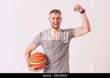 Attraktiver bärtiger Mann, der den Arm hochgehoben und lächelt, während er auf weißem Hintergrund isoliert ist. Junger Flusspferd, der den Sieg feiert und Basketballbal hält Stockfoto