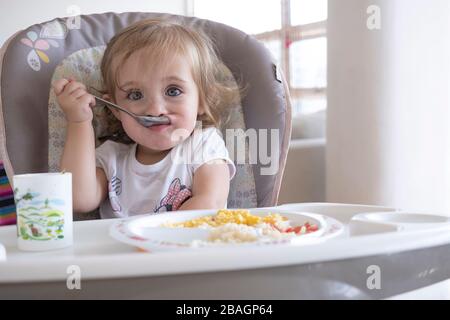 Kleines Mädchen, das allein auf dem Stuhl am Tisch isst. Stockfoto