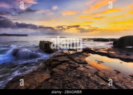 Sandsteinplateau bei Ebbe mit kleiner Pfütze aus Meerwasser, das gelbe Wolken bei Sonnenaufgang an den Nordstränden von Sydney reflektiert. Stockfoto