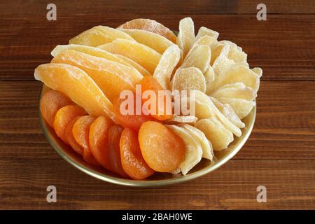 Scheiben aus kandierten Ingwerwurzeln, Stücke getrockneter Mango-Früchte und getrocknete Aprikosen, die auf einer goldenen Platte auf einem Holztisch liegen. Nahaufnahme. Makro Stockfoto