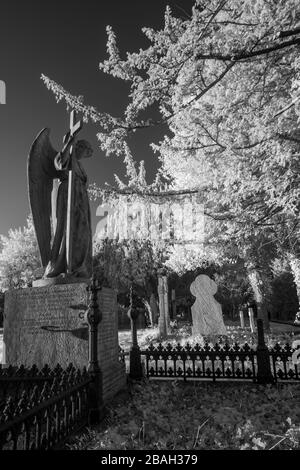 Eine Statue eines Engels, der ein Kreuz hält, blickt über mehrere Gräber auf einem Friedhof. Im Nahinfrarotbereich (720 nm) aufgenommen. Stockfoto