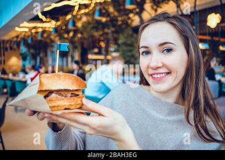 Mädchen hält einen Hamburger auf dem Hintergrund des Restaurants und schaut auf die Kamera. Nahaufnahme. Stockfoto