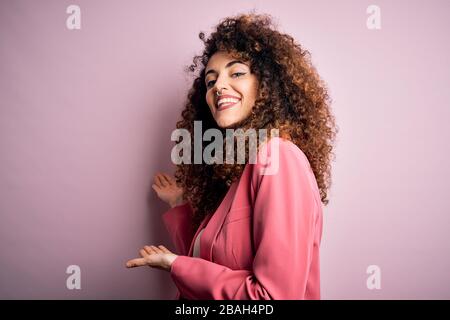 Junge, schöne Geschäftsfrau mit lockigen Haaren und durchstechenden, eleganten Blazer, die dazu einladen, mit offener Hand in lächelnde Natur zu treten Stockfoto