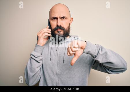 Gutaussehender kahler Mann mit Bart, der auf dem Smartphone mit verärgertem Gesicht spricht, negatives Zeichen, das mit Daumen nach unten ablehnt Stockfoto