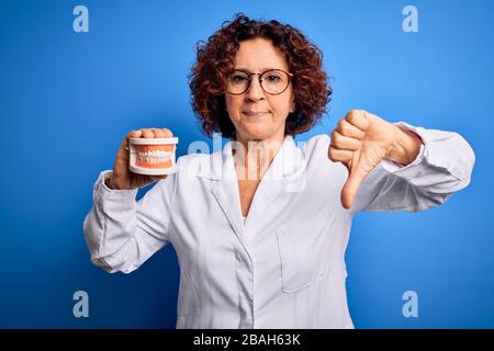 Zahnärztin im mittleren Alter, die einen Mantel trägt, der die Zähne aus Kunststoff über blauem Hintergrund mit wütenden Gesichtern hält, ein negatives Zeichen, das mit den Daumen ablehnt Stockfoto